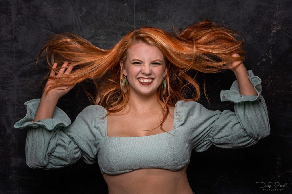 lady in a green crop top smiling widely with both hands on bright orange hair 