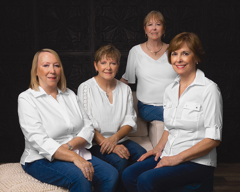 four elderly women in white tops and blue jeans sitting on a couch in a studio
